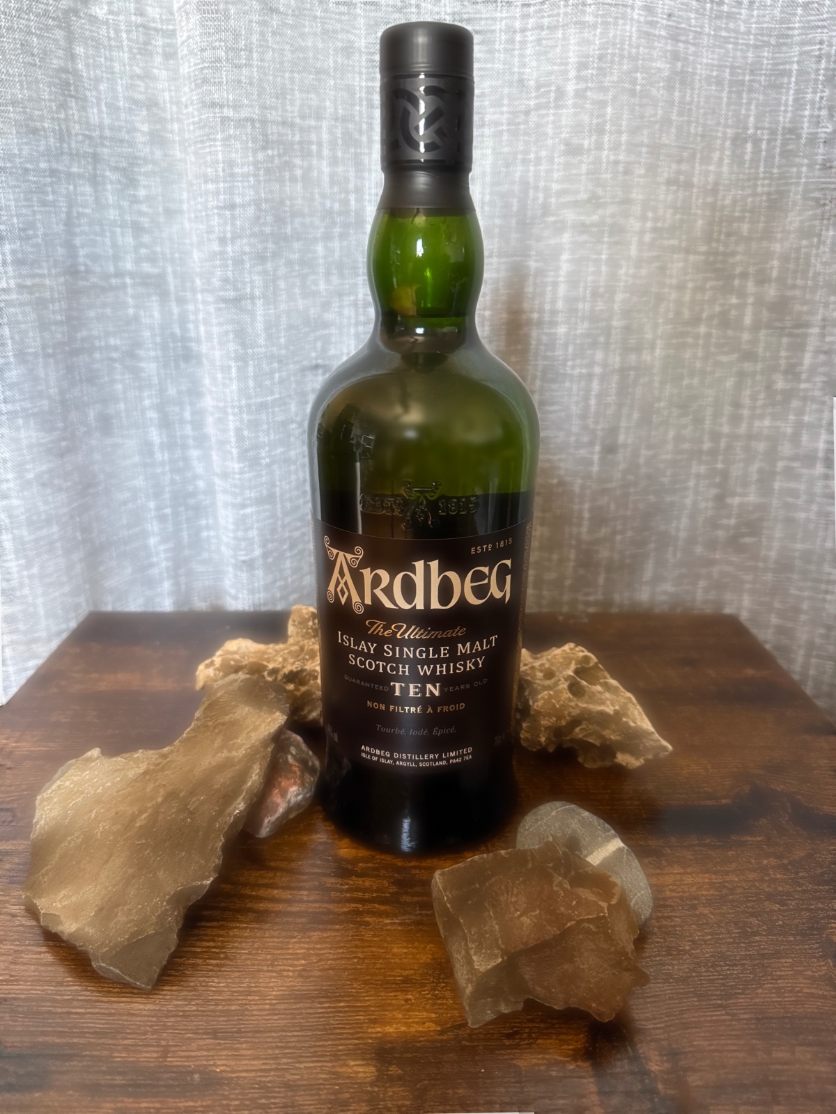 Ardbeg Ten Islay single malt bottle on dark wood, flanked by pale translucent quartz stones against a soft linen backdrop
