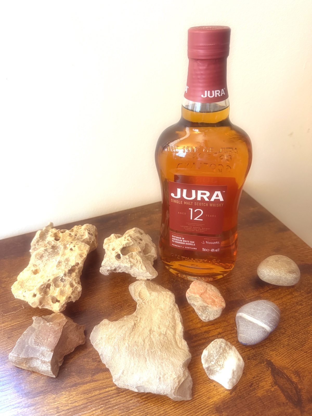 Jura 12 Year Old single malt bottle on dark wood, surrounded by pale fossil-bearing stones and smooth beach pebbles